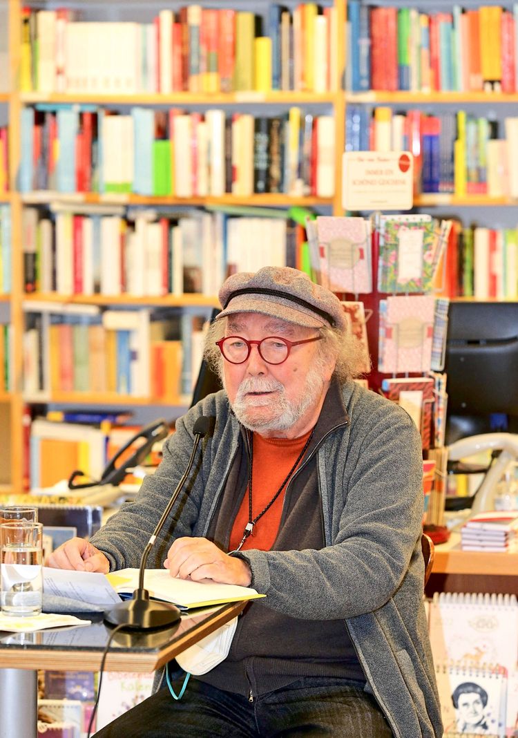 Ein älterer Mann mit Brille und Schirmmütze sitzt an einem Tisch in einer Buchhandlung. Vor ihm liegen ein Mikrofon, ein Glas Wasser und ein aufgeschlagenes Buch. Im Hintergrund sind Bücherregale zu sehen.