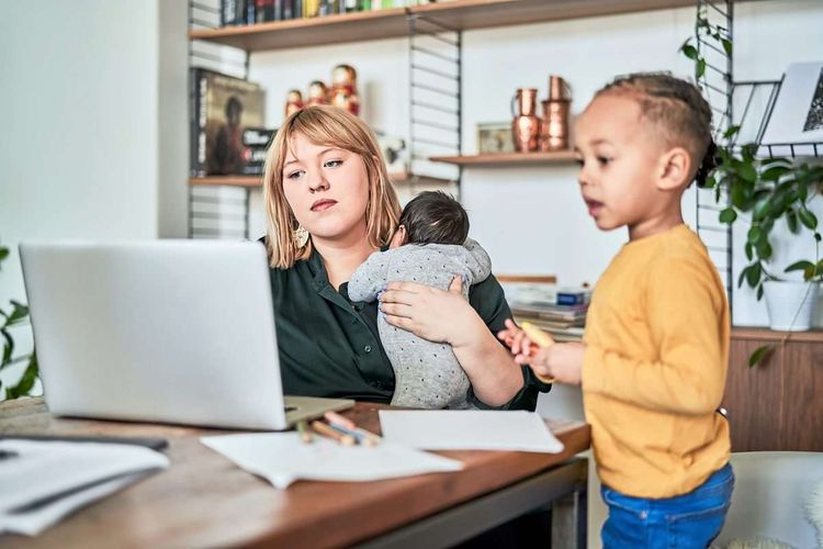 Eine Mutter mit Baby am Arm sitzt vor dem Laptop, daneben steht ein kleiner Junge