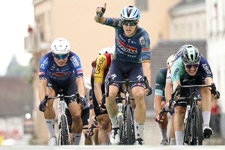 Ein Radrennfahrer im Trikot des Teams Soudal Quick-Step hebt jubelnd eine Hand, während er als Erster die Ziellinie überquert. Neben ihm kämpfen weitere Fahrer, darunter einer im blauen und ein anderer im mintgrünen Trikot, um die vorderen Plätze. Im Hintergrund verschwommene Architektur.