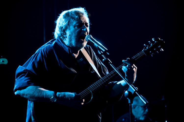 Ein Mann mit grauen Haaren spielt Gitarre und singt in ein Mikrofon während eines Konzerts, das in einer dunklen Bühnenbeleuchtung stattfindet. Im Hintergrund ist ein Musiker an einem Schlagzeug zu erkennen.