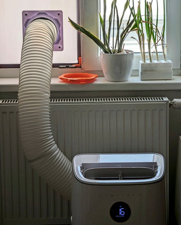 Eine mobile Klimaanlage mit einem Schlauch, der durch eine Fensterdichtung nach draußen führt. Auf der Fensterbank stehen zwei Pflanzen in weißen Gefäßen neben einem orangefarbenen Gegenstand. Das Gerät zeigt eine Temperatur von 16 Grad auf der Anzeige.