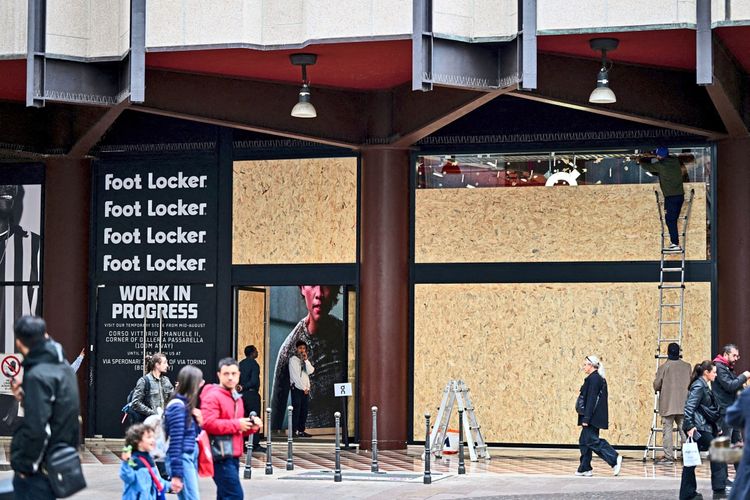 Eine Person steht auf einer Leiter und befestigt Holzplatten vor der Fassade eines Foot Locker-Geschäfts. Fußgänger passieren den Bereich auf einer Straße. Das Geschäft ist teilweise mit Sperrholz verkleidet.