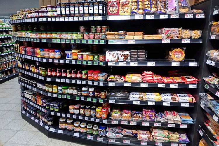 Ein Supermarktregal mit verschiedenen Lebensmitteln wie Marmelade, Brotaufstrichen, Gebäck und Nutella. Die Produkte sind in mehreren Reihen ordentlich sortiert, mit Preisschildern versehen.