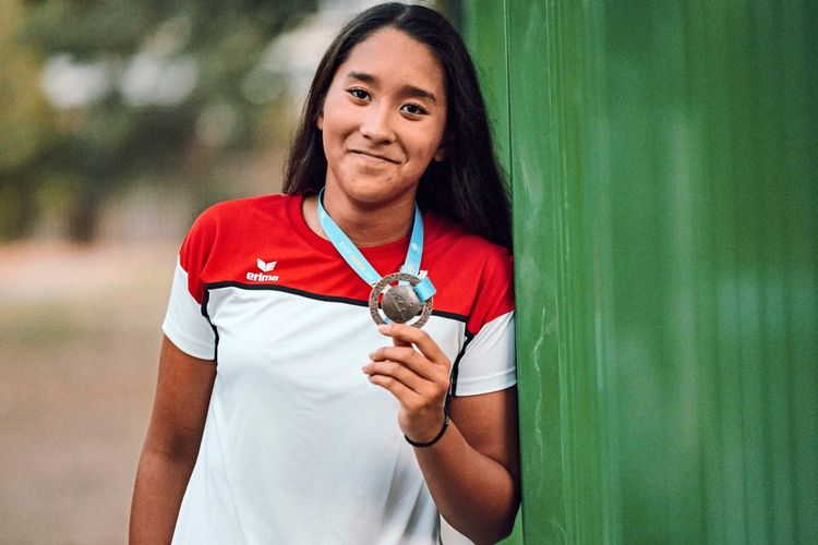 Eine junge Frau in einem rot-weißen T-Shirt hält eine Medaille in der Hand und lächelt in die Kamera. Sie steht an einer grünen Wand, im Hintergrund ist unscharfe Natur zu erkennen.