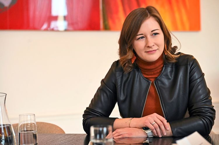 Eine Frau mit schulterlangem braunem Haar sitzt an einem Tisch. Sie trägt einen orangefarbenen Rollkragenpullover, eine schwarze Lederjacke und orangefarbene Ohrringe. Vor ihr befinden sich Gläser und eine Karaffe. Im Hintergrund ist ein modernes Kunstwerk in Rot- und Orangetönen zu sehen.