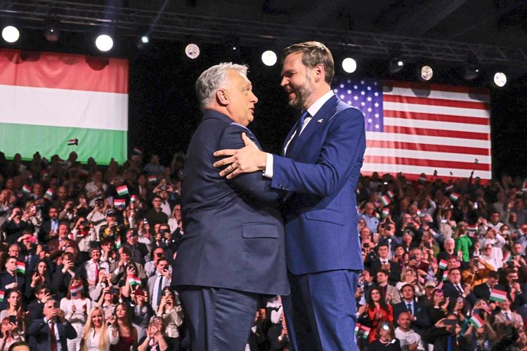 US-Vizepräsident J. D. Vance und Ungarns Premier Viktor Orbán schütteln sich auf einer Bühne die Hand. Im Hintergrund ist eine Menschenmenge zu sehen, die kleine ungarische Fähnchen schwenkt. Links befindet sich eine große ungarische Flagge, rechts eine US-amerikanische Flagge.