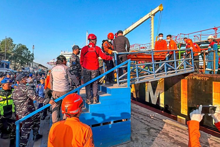Rettungskräfte transportieren einen Verletzten auf einer Trage von einem Schiff. Eine Menschenmenge, darunter Sicherheits- und Rettungspersonal in Uniform und Schutzkleidung, steht an einem Hafen, während die Rettungsaktion fortgeführt wird. Im Hintergrund sind ein Schlepper und Hafeneinrichtungen zu sehen.