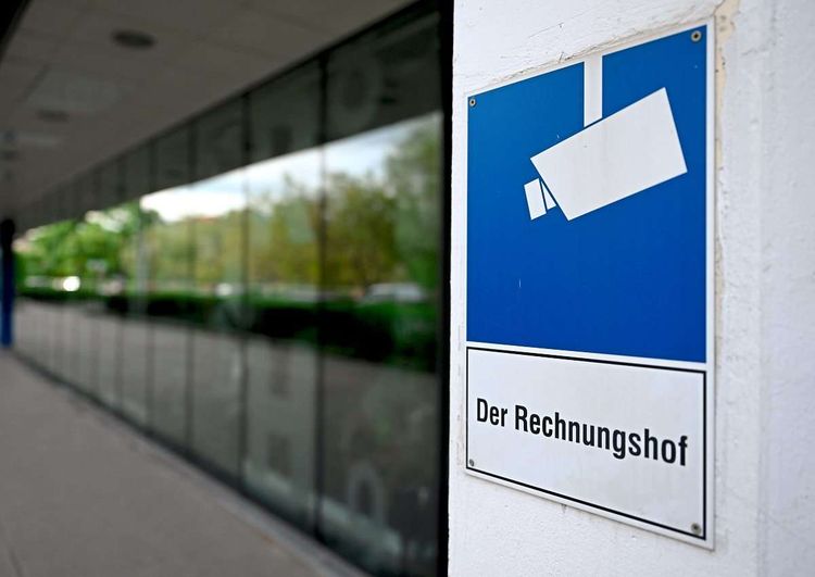 Auf einem weißen Schild steht Rechnungshof. Darüber ist ein weißes Kamerasymbol auf blauem Hintergrund montiert. Links daneben die Glasfront eines Gebäudes.