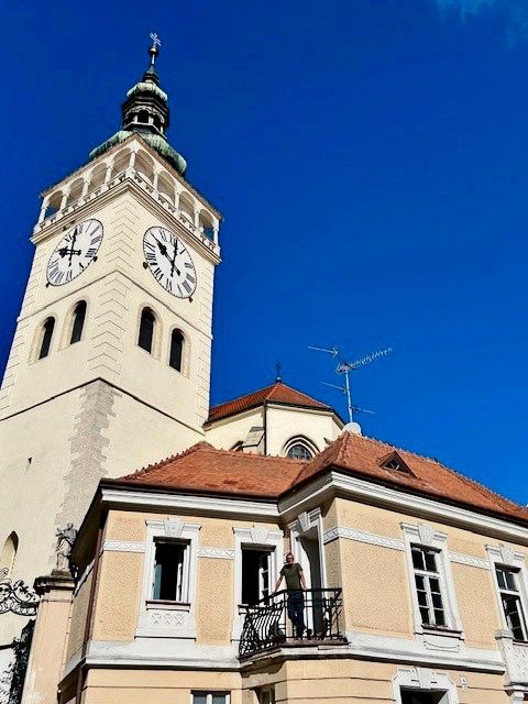 Ein großes Gebäude mit einem Turm und einer Uhr, umgeben von strahlend blauem Himmel. Eine Person steht auf einem kleinen Balkon eines Gebäudes mit roten Ziegeldächern und hellen Fassaden.