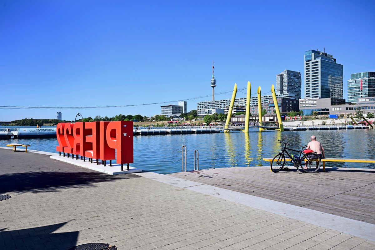 Ein Bild zeigt eine Uferpromenade mit einem großen roten Schriftzug „PIER22“. Im Hintergrund sind ein Wasserbecken, moderne Gebäude, ein gelber Kunstinstallationsbogen und ein Fernsehturm zu sehen. Im Vordergrund sitzt eine Person ohne Oberbekleidung auf einer Bank neben einem Fahrrad, während die Umgebung sonnig und klar ist.