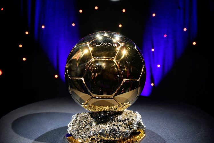 Die goldene Ballon-d'Or-Trophäe auf einem dunklen Podest mit lila und roten Lichtpunkten im Hintergrund.