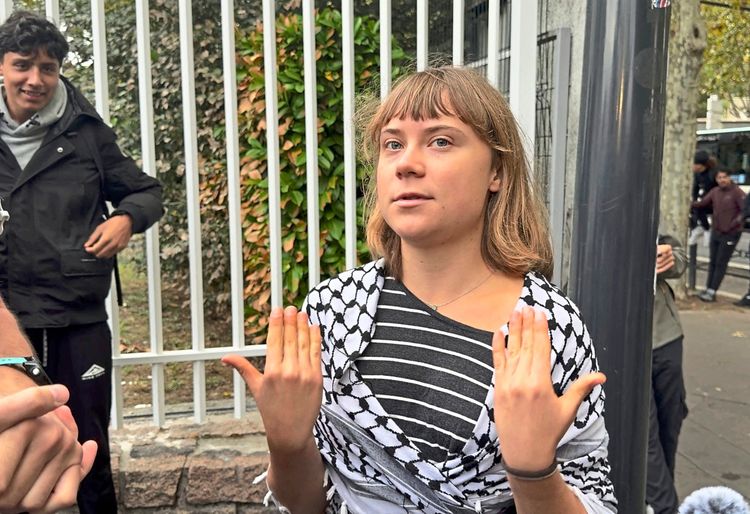 Eine Frau mit blonden Haaren steht vor einem Zaun. Sie trägt ein gestreiftes Oberteil und ein Tuch mit schwarz-weißem Muster um die Schultern. Ihre Hände sind erhoben, und sie blickt ernst. Im Hintergrund sind weitere Personen und Bäume zu sehen.