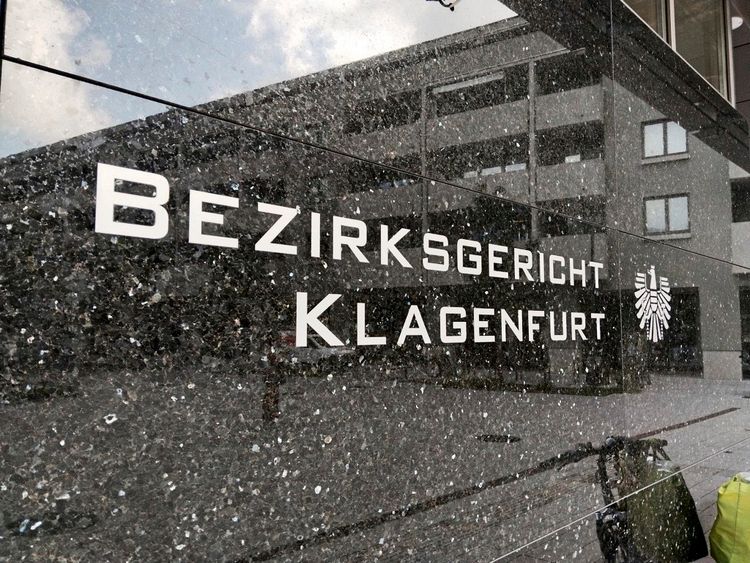 Außenansicht einer glänzenden Fassade mit der Aufschrift „Bezirksgericht Klagenfurt“ und einem Wappen rechts daneben. Im Hintergrund spiegelt sich ein Gebäude.