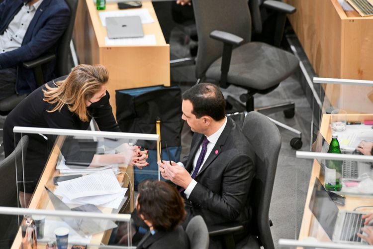 Eine Frau und ein Mann sprechen miteinander in einem Bürobereich, der durch Plexiglasscheiben abgetrennt ist. Auf dem Tisch liegen Dokumente, und im Hintergrund sind weitere Personen sowie Laptops und Bürostühle sichtbar.
