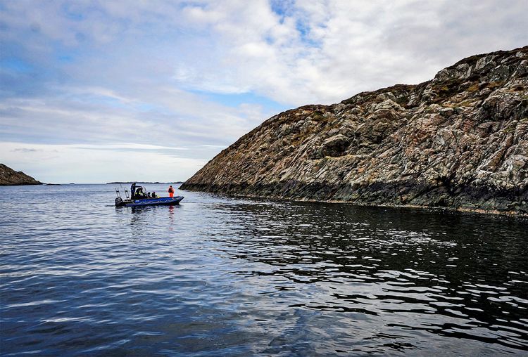 Eine felsige Insel mit steil abfallenden Klippen ins Meer, Hespriholmen, umgeben von ruhigem Wasser. Ein kleines Boot mit mehreren Personen ist nahe der Küste zu sehen, während der Himmel teilweise bewölkt ist.