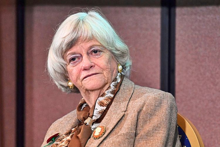 Eine ältere Frau mit grauem Haar sitzt vor einer neutralen, braunen Hintergrundwand. Sie trägt einen beigen Blazer, ein gemustertes Halstuch und Ohrringe.