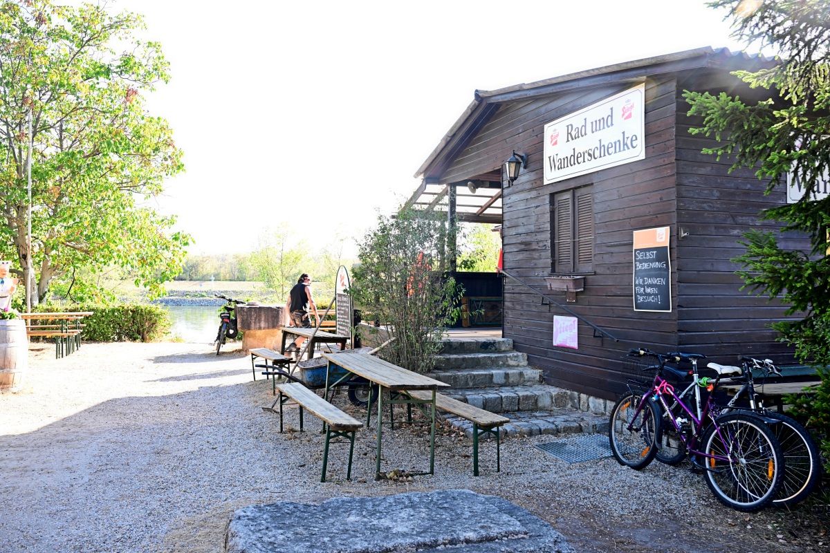 Ein kleines Holzhäuschen mit der Aufschrift „Rad und Wanderschenke“. Es befinden sich Holzbänke und -tische im Außenbereich. Zwei Fahrräder stehen rechts. Ein Weg führt in Richtung eines Sees im Hintergrund, umgeben von grünen Bäumen und sonniger Landschaft.