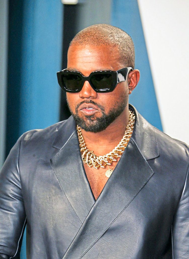 Kanye West auf dem roten Teppich einer Preisverleihung. Er trägt ein Ledersakko, Goldschmuck und dunkle Sonnenbrillen.