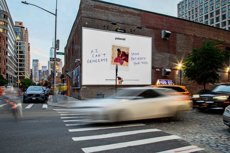 Eine Straßenszene in einer Stadt mit einem großen Werbeplakat auf einer Backsteinwand. Auf dem Plakat steht: „A.I. CAN’T GENERATE SAND BETWEEN YOUR TOES“ und zeigt ein Polaroid-Bild einer Person im Sand. Unten steht der Slogan von Polaroid. Autos und Fußgänger sind in Bewegung, die Umgebung zeigt moderne Gebäude im Hintergrund.
