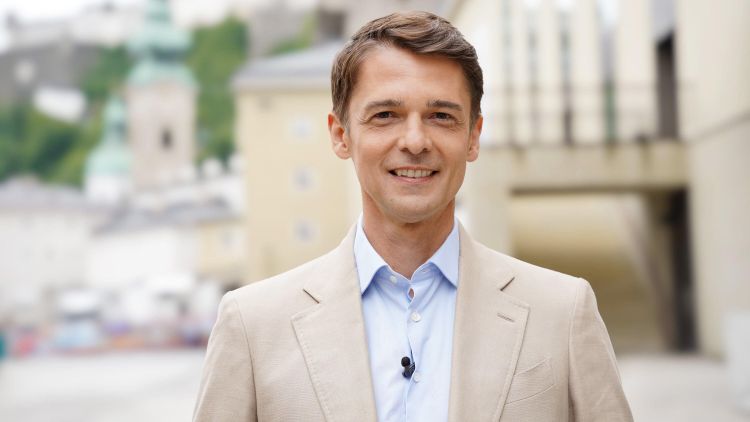 Mann mit brünettem Scheitel, hellem Sakko, hellblauem Hemd lächelt in die Kamera.