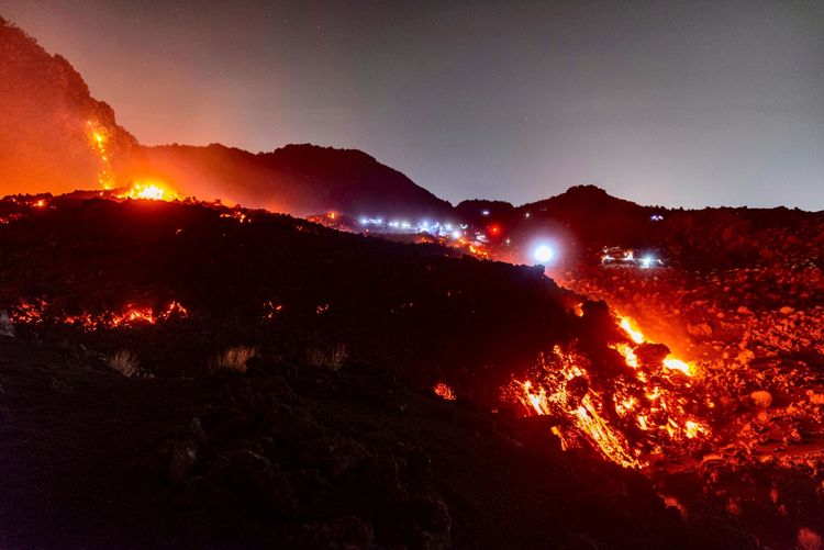 ALT-Text: Nachtsicht auf den Ausbruch des Vulkans Ätna in Catania, Italien, mit leuchtenden Lavaströmen, die aus aktiven Entlüftungen fließen. Die Umgebung ist von glühender Lava beleuchtet, während im Hintergrund Nachthimmel und Vulkanlandschaften sichtbar sind.