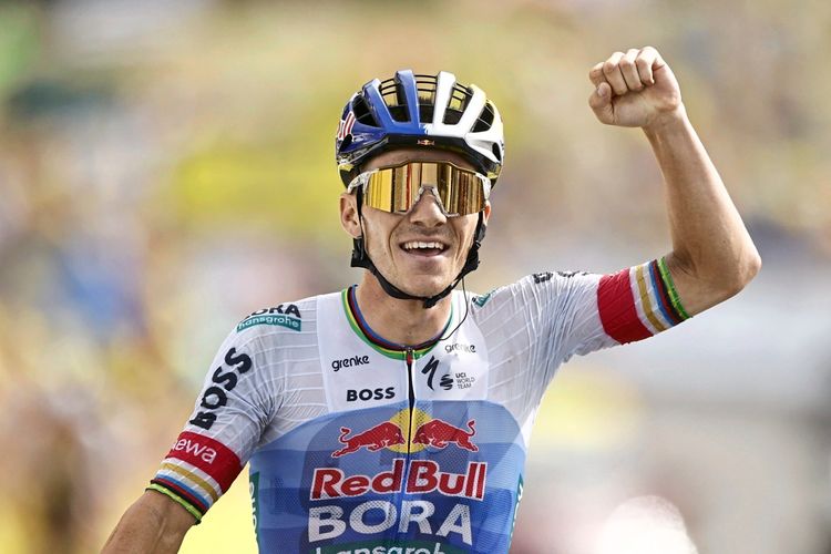 Ein Radrennfahrer im Trikot des Red Bull - Bora - Hansgrohe-Teams hebt triumphierend die Faust, während er die Ziellinie überquert. Er trägt einen Helm und eine Sonnenbrille. Im Hintergrund ist die verschwommene Kulisse eines Rennens zu erkennen.