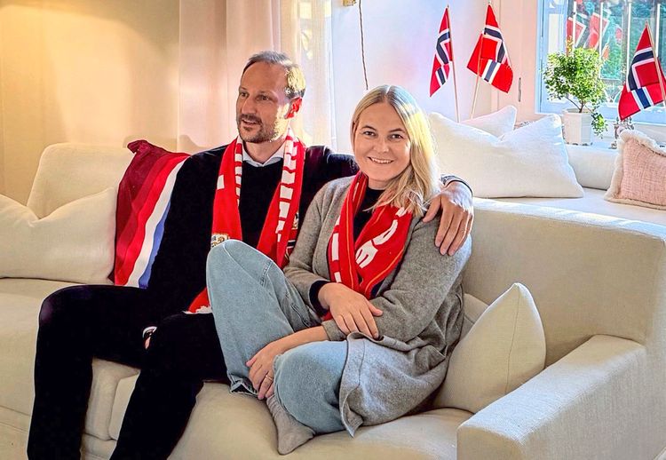 Ein Mann und eine Frau sitzen gemeinsam auf einem hellen Sofa in einem Wohnzimmer. Sie lächeln und tragen rote Schals mit Fußballmotiven. Im Hintergrund sind norwegische Flaggen und ein Fenster mit einfallendem Tageslicht zu sehen. Die Szene wirkt gemütlich und feierlich.