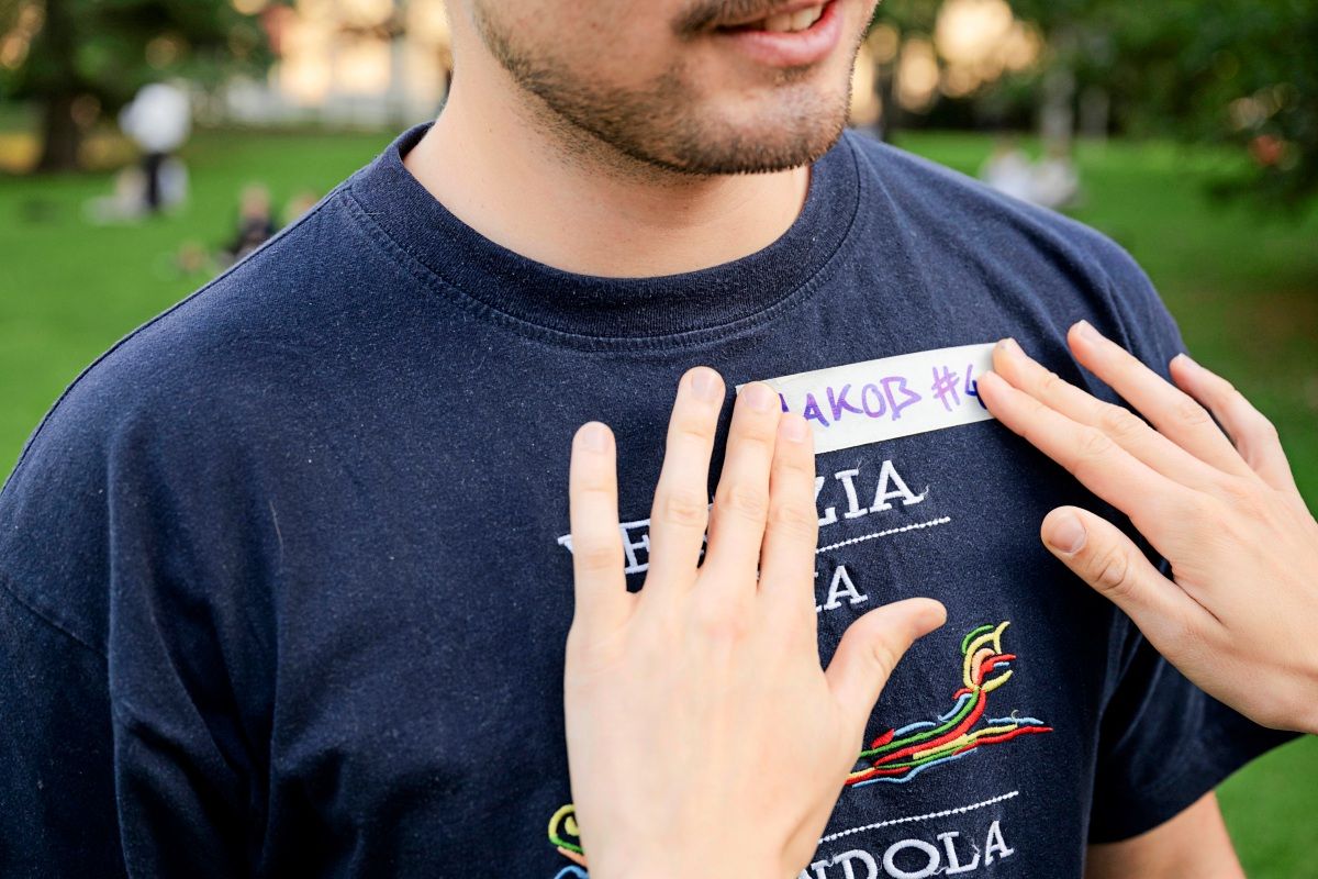Ein Mann trägt ein dunkelblaues T-Shirt, während Hände ein Namensschild mit handgeschriebenem Text auf seine Brust kleben. Hintergrund zeigt eine grüne Parklandschaft.