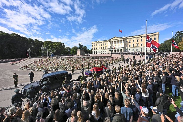 Eine große Menschenmenge versammelt sich vor dem Königspalast in Oslo, um die royale Prozession zu sehen, bei der der mit einer roten Flagge bedeckte Sarg von König Harald V. von Norwegen vorbeigeführt wird. Soldaten in Uniform flankieren die Szene, während norwegische Flaggen auf Halbmast wehen. Im Hintergrund ist der Palast mit einer Statue und einem mit Blumen bedeckten Platz sichtbar.
