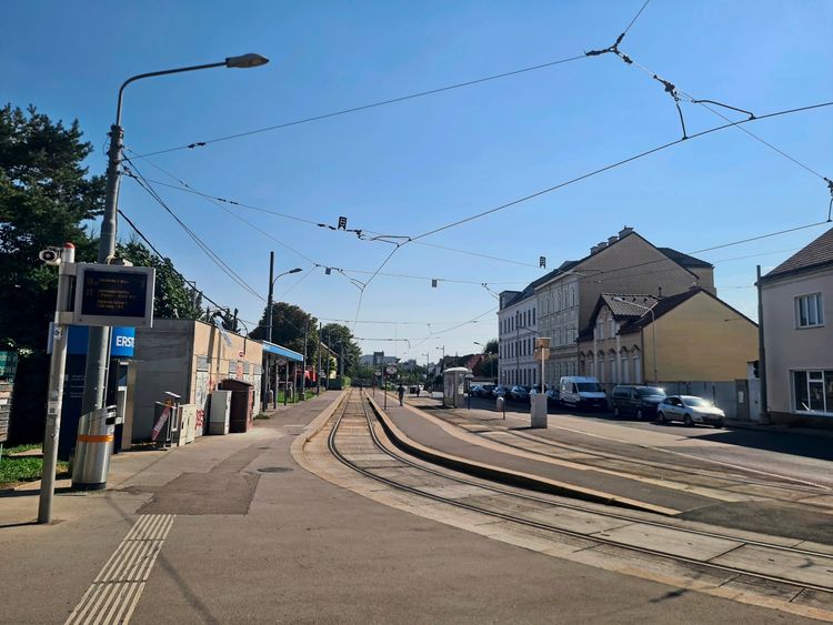 Straßenbahnhaltestelle an einer Straße bei klarem, sonnigem Himmel. Schienen verlaufen durch die Haltestelle. Auf der linken Seite ein Fahrplananzeiger, ein Bankautomat und kleine Geschäfte. Rechts sind Gebäude und parkende Autos zu sehen. Oberleitungen für die Straßenbahn verlaufen über die Straße.