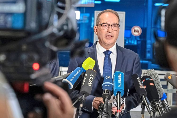 Ein Mann in Anzug und Krawatte spricht bei einer Pressekonferenz. Vor ihm sind mehrere Mikrofone von verschiedenen Medienhäusern sichtbar. Der Hintergrund zeigt einen Raum mit Büro- oder Kontrollraumeinrichtung in kühlem, bläulichem Licht.