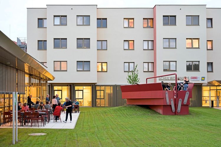 Ein moderner Gebäude-Komplex mit heller Fassade. Im Vordergrund eine rote, kunstvolle Skulptur in Form eines Werkzeugkastens von Anna Vasof. Links sitzen Menschen an Tischen im Außenbereich, beleuchtet durch warmes Licht aus dem Gebäude. Im Hintergrund Rasenfläche und Fenster des Wohnhauses.