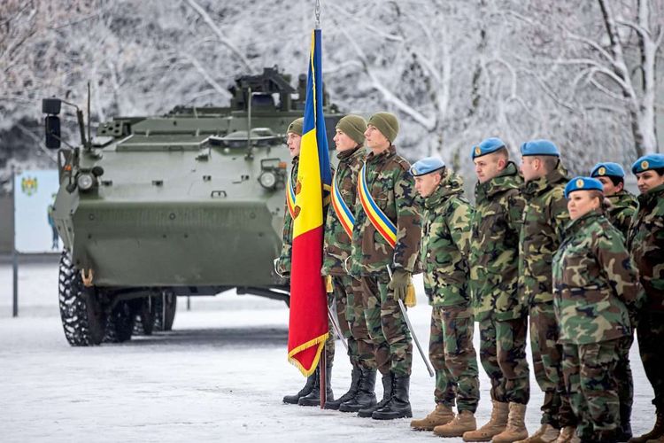 Angehörige der Streitkräfte stehen in Uniform und mit Fahne auf einem verschneiten Platz. Im Hintergrund links ist ein Radschützenpanzer 