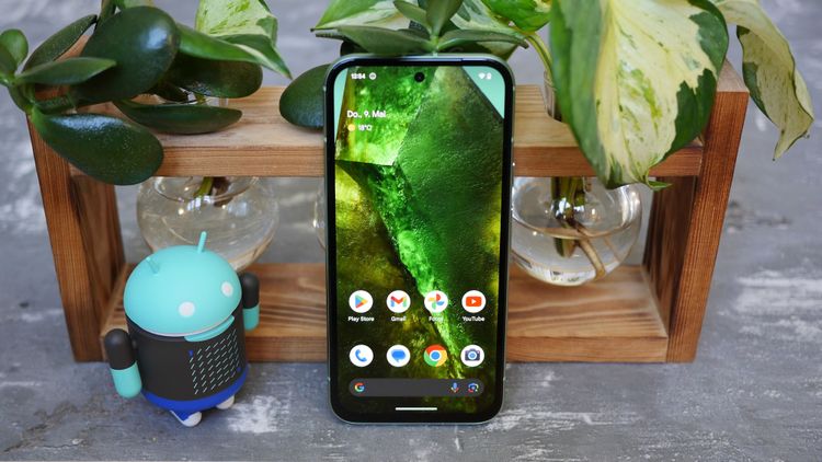 Das Pixel 8a mit aktiviertem Bildschirm, zu sehen ist der Homescreen. Daneben steht eine Android-Figur, das Smartphone ist an eine Pflanzenhalterung gelehnt, sind im  Hintergrund zu sehen