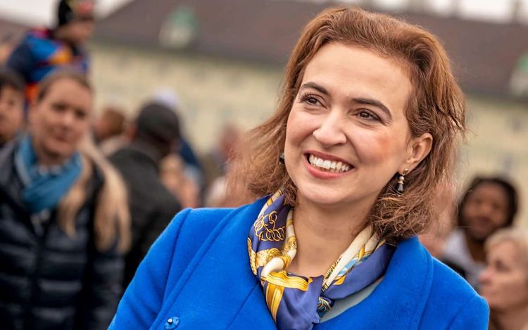 Alma Zadic in blauem Mantel vor Menschenmenge, lächelnd.