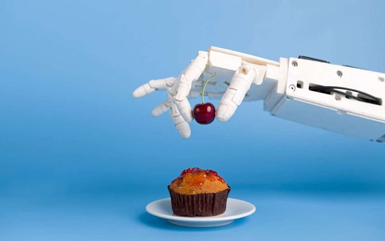 Roboterhand legt Kirsche auf Muffin.
