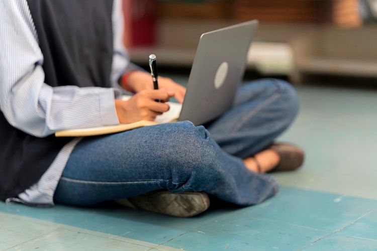 Eine Person sitzt auf einem Boden, trägt Jeans und ein Hemd und benutzt einen Laptop, während sie in ein Notizbuch schreibt.