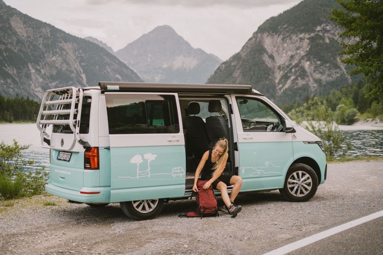 Frau mit Wanderschuhen und Wanderrucksack sitzt in der Türöffnunge eines Campingbuses, der auf dem Parkplatz vor einem See mit Berpanorama steht.