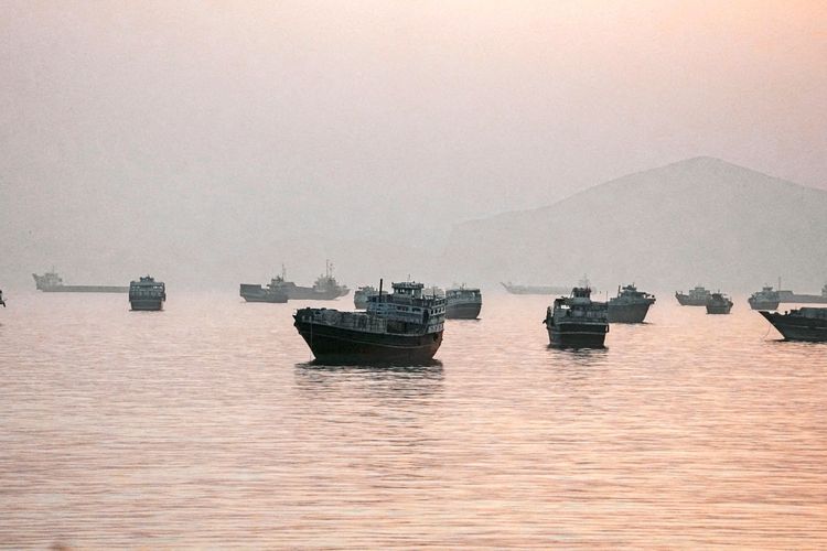 Schiffe liegen vor Anker in der Straße von Hormus, mit leichtem Dunst und einer Bergsilhouette im Hintergrund bei Sonnenuntergang, gesehen von Musandam, Oman.
