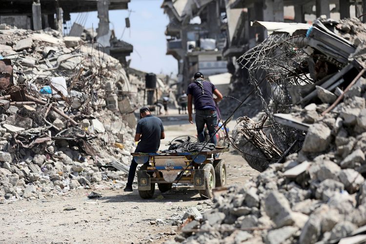 Zwei Personen schieben einen beladenen Karren durch eine Straße, die von Trümmern und zerstörten Gebäuden gesäumt ist, in der Nähe des Jabalia-Lagers im nördlichen Gaza-Streifen. Die Szene zeigt die Auswirkungen von Zerstörung und schwierigen Lebensbedingungen in der Region.