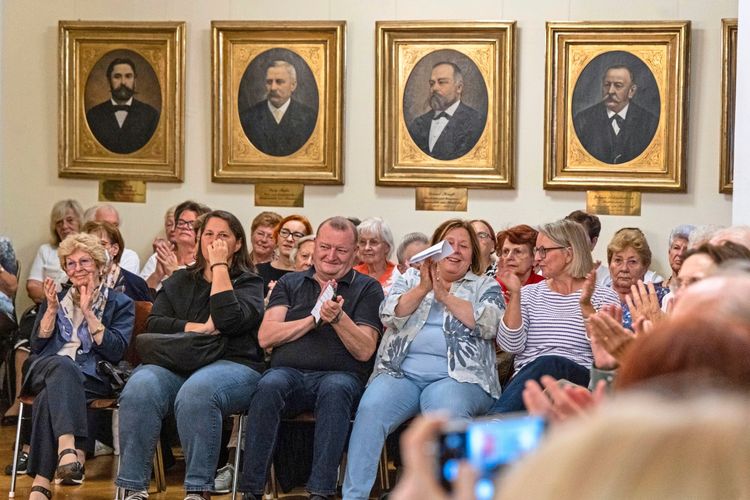 Eine Gruppe von Zuschauern sitzt in einem Raum mit Porträts an der Wand und applaudiert. Die Atmosphäre wirkt aufmerksam und respektvoll, vermutlich bei einer Gedenkveranstaltung oder kulturellen Veranstaltung.
