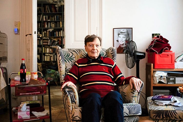 Ein älterer Mann sitzt in einem gepolsterten Sessel in einem Wohnzimmer. Er trägt einen gestreiften Pullover und blickt direkt in die Kamera. Im Hintergrund sind ein Bücherregal, eine Tür und eine Wand mit Fotos und Gegenständen zu sehen. Auf einem kleinen Tisch neben ihm stehen einige Alltagsgegenstände.