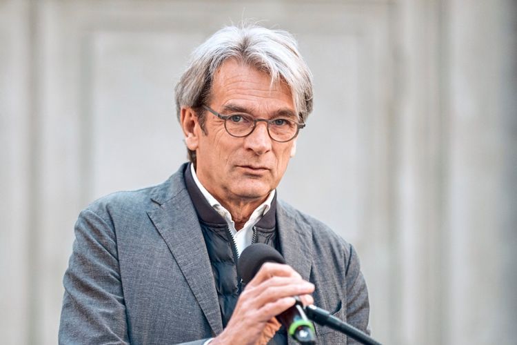 Ein Mann mit grauem Haar und Brille spricht in ein Mikrofon. Er trägt ein helles Hemd und ein dunkles Sakko. Der Hintergrund ist neutral und unscharf.