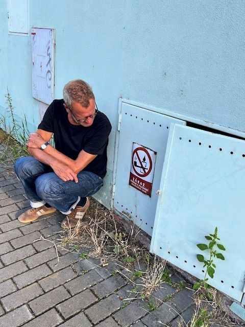 Der Autor und Künstler Tex Rubinowitz in Jeans und schwarzem T-Shirt hockt neben einer hellblauen Wand mit einer Türklappe. Auf der Klappe befindet sich ein Schild mit einem Rauchen-verboten-Symbol. Der Boden ist mit Betonsteinen gepflastert, und daneben wächst etwas Gras.