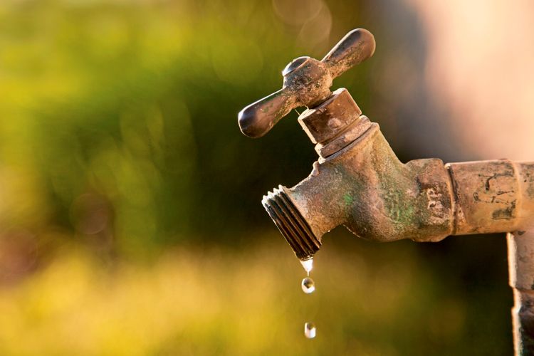Rostiger Außenwasserhahn, aus dem langsam Wassertropfen fallen, vor einem unscharfen, grün-gelben Hintergrund.