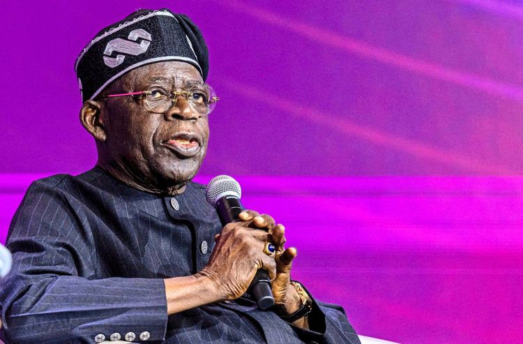 Ein Mann in traditioneller nigerianischer Kleidung und Mütze hält ein Mikrofon und spricht während einer Podiumsdiskussion. Der Hintergrund ist in lila Farbtönen gehalten.