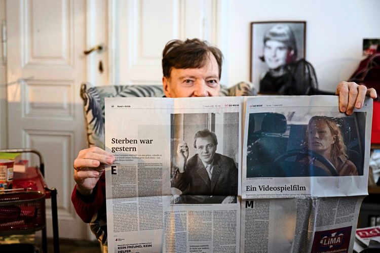 Ein Mann sitzt in einem Raum mit einem retro-dekorierten Hintergrund und hält eine Zeitung in die Kamera. Die Zeitung enthält Artikel mit den Überschriften „Sterben war gestern“ und „Ein Videospiel­film.“ Im Hintergrund ist ein gerahmtes Porträt sowie Möbelstücke zu sehen.