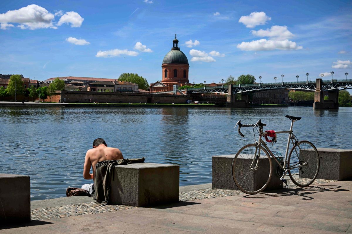 Ein Mann sitzt mit freiem Oberkörper am Ufer der Garonne in Toulouse, Frankreich, mit Blick auf die Chapelle Saint-Joseph de la Grave. Neben ihm ist ein Fahrrad abgestellt.