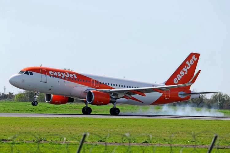 Ein EasyJet Airbus A320 beim Landen auf der Landebahn des Flughafens Orly. Rauch ist sichtbar an den Rädern, die den Boden berühren, und das Flugzeug trägt eine orange-weiße Lackierung. Im Hintergrund sind grasbewachsene Flächen und Bäume zu sehen.