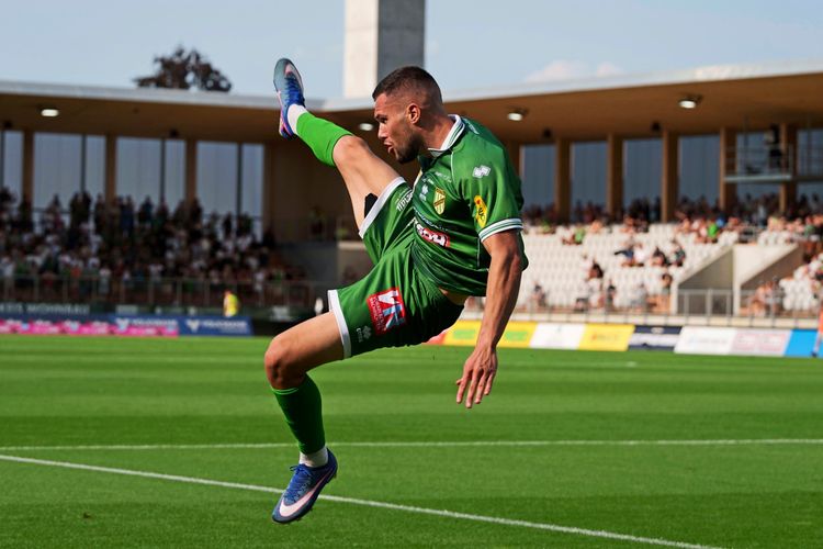 Ein Fußballspieler in grünem Trikot des SC Austria Lustenau springt mit gestrecktem Bein in die Luft während eines Spiels. Im Hintergrund sind Zuschauer auf einer Tribüne zu sehen.